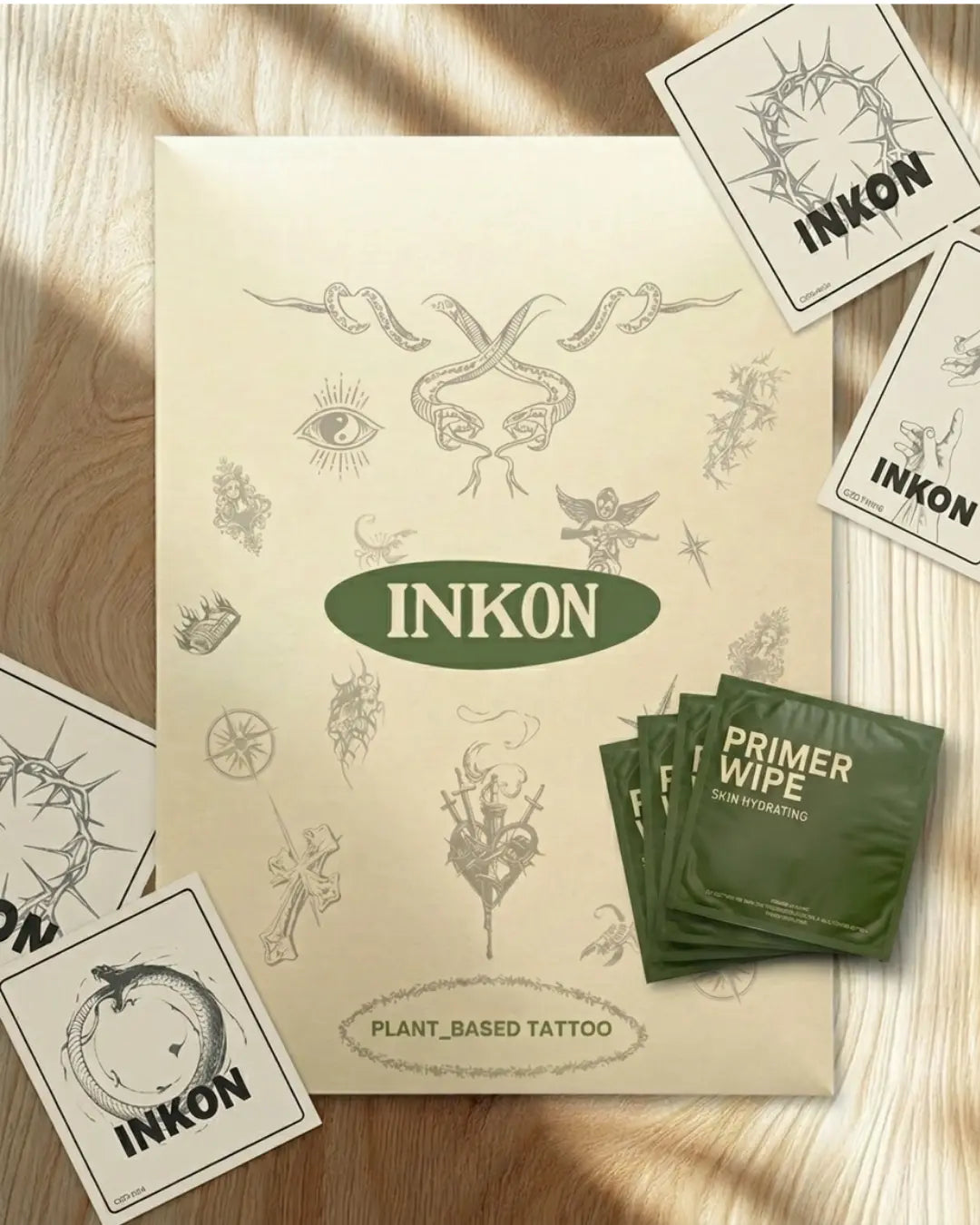 Kit de empaque completo INKON Tattoos For Now sobre una superficie de mármol – sobre verde de la marca con ilustraciones florales y de mariposas, sachet humectante para preparar la piel, tarjeta con instrucciones de aplicación con código QR y sticker de la marca INKON Tattoo